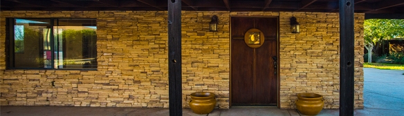 Front door to a southwest style home. House has a stone veneer, a wood door with a window, and two lamps on either side of the door. There is a window to the left of the door. The house has a cement sidewalk entryway covered by a wood arbor. Photo by Emma Rosenthal